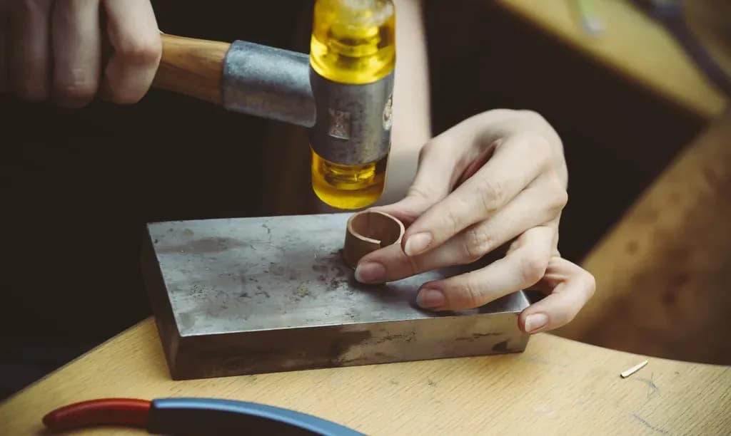A jeweller hand-forming a silver ring with a mallet on a steel bench block.