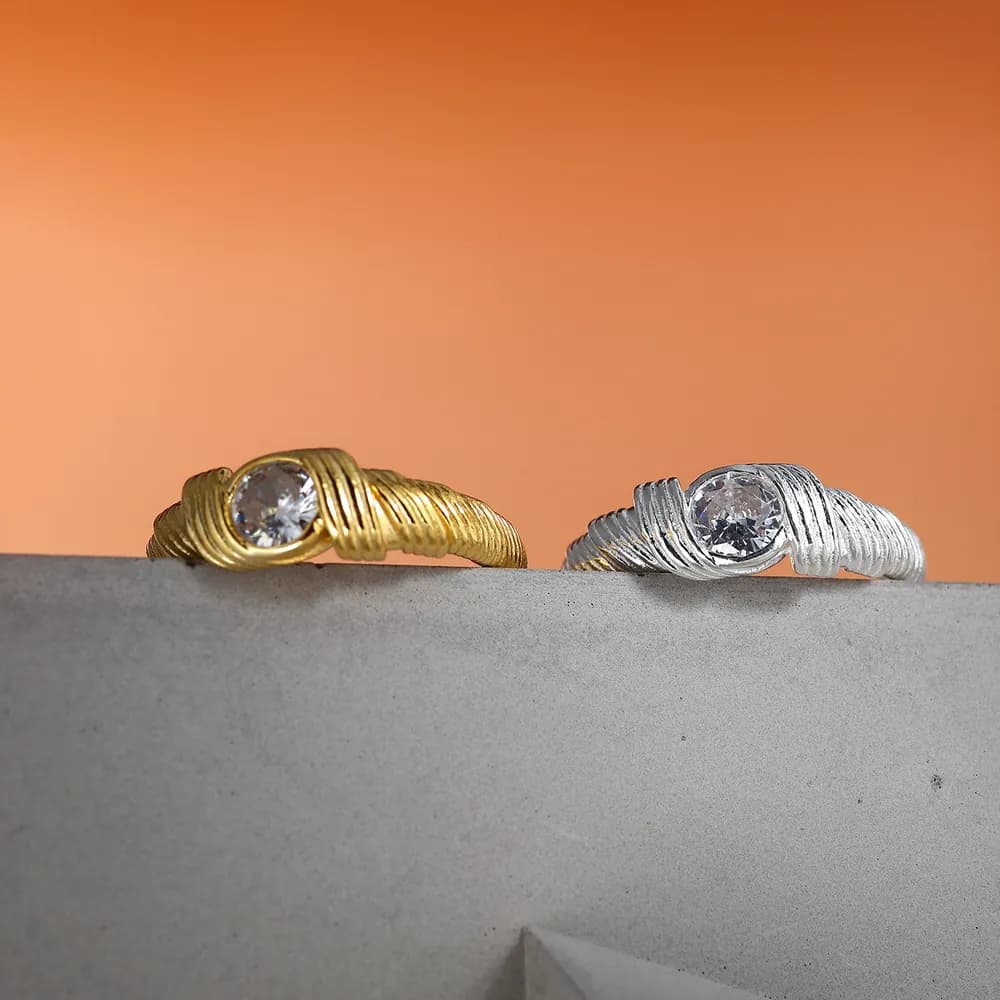 Two twisted ribbon rings, one gold-tone and one silver, with bezel-set round stones resting on a concrete ledge.