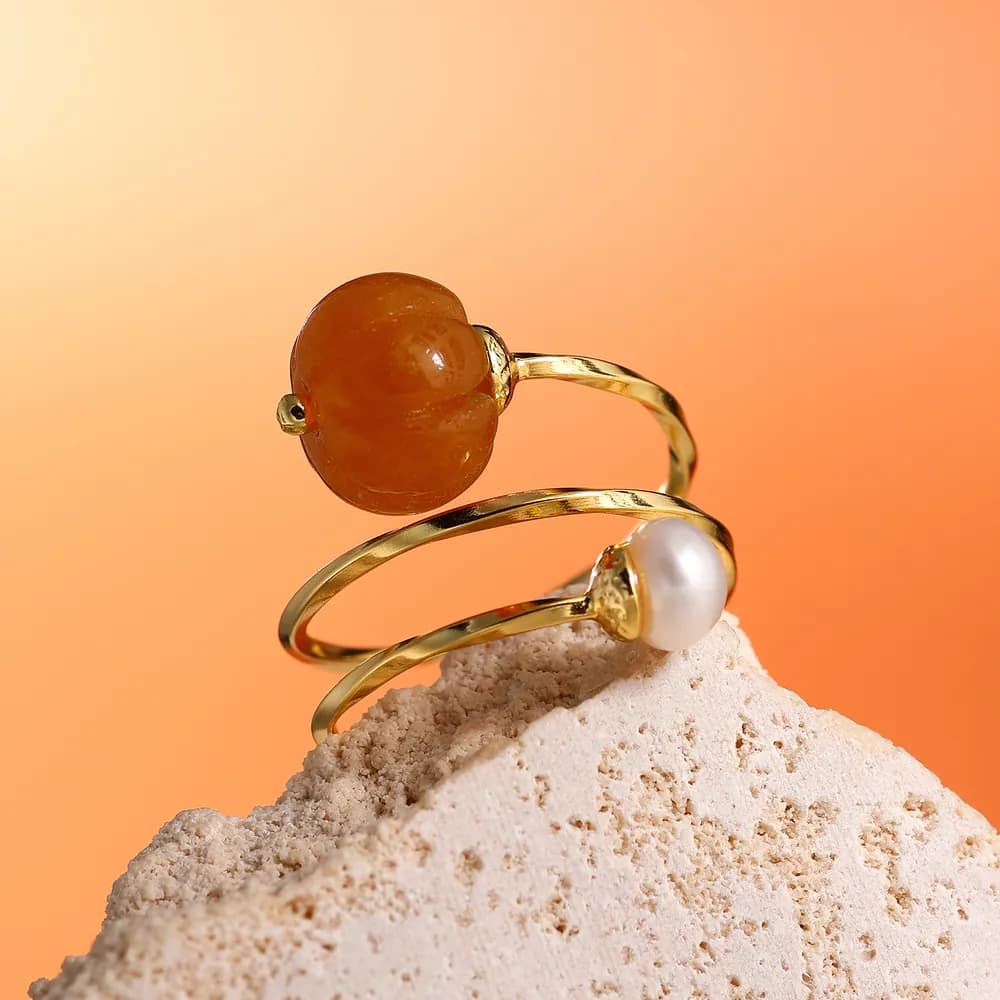 Two thin polished rings stacked beside a textured rock, one with an orange carnelian cabochon and the other with a white pearl, against an orange background.