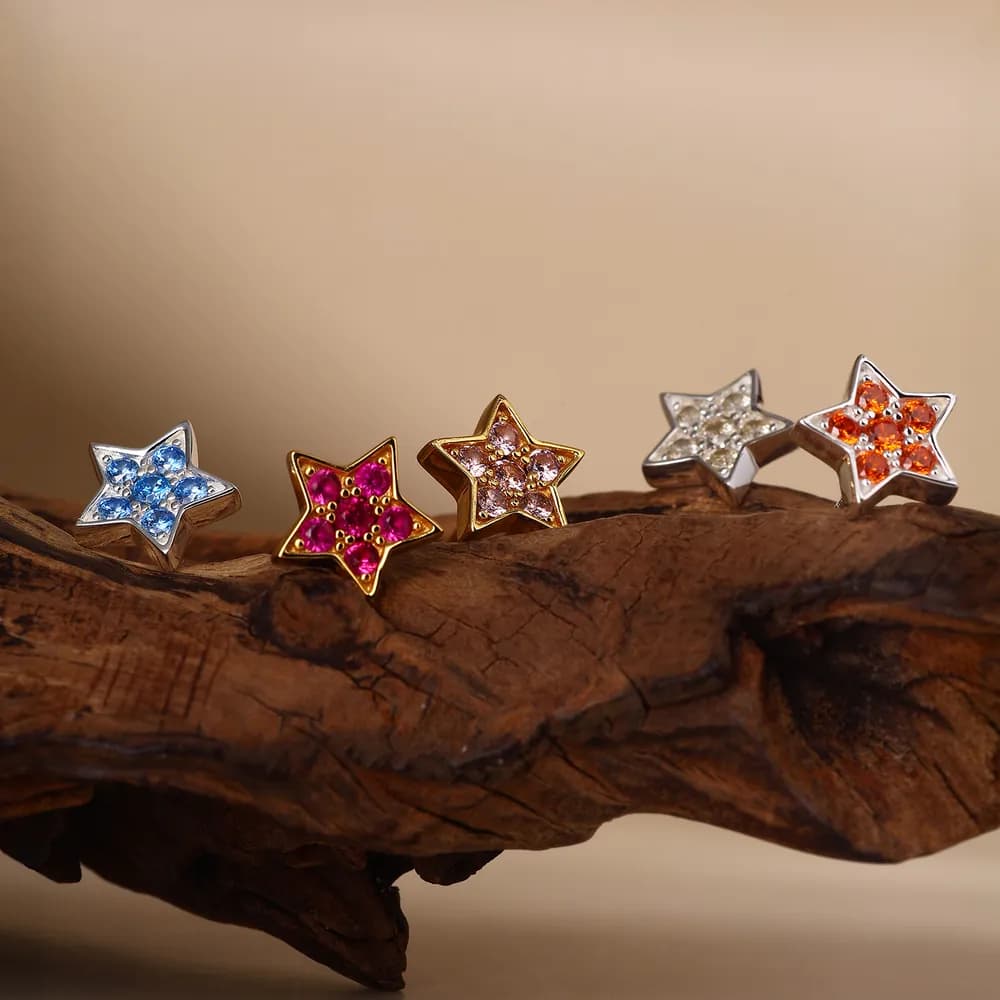 Five star-shaped pavé earrings in silver and warm-tone settings with colorful stones arranged on a wooden surface.