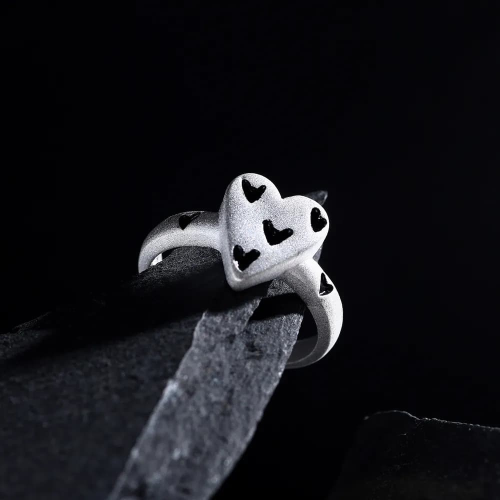 A frosted silver ring with a heart-shaped top adorned with small black enamel hearts sits on a dark, textured surface.