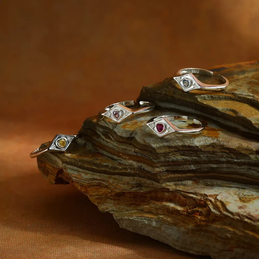 Five sterling silver rings with diamond-shaped settings and heart-shaped enamel centers resting on a layered rock.