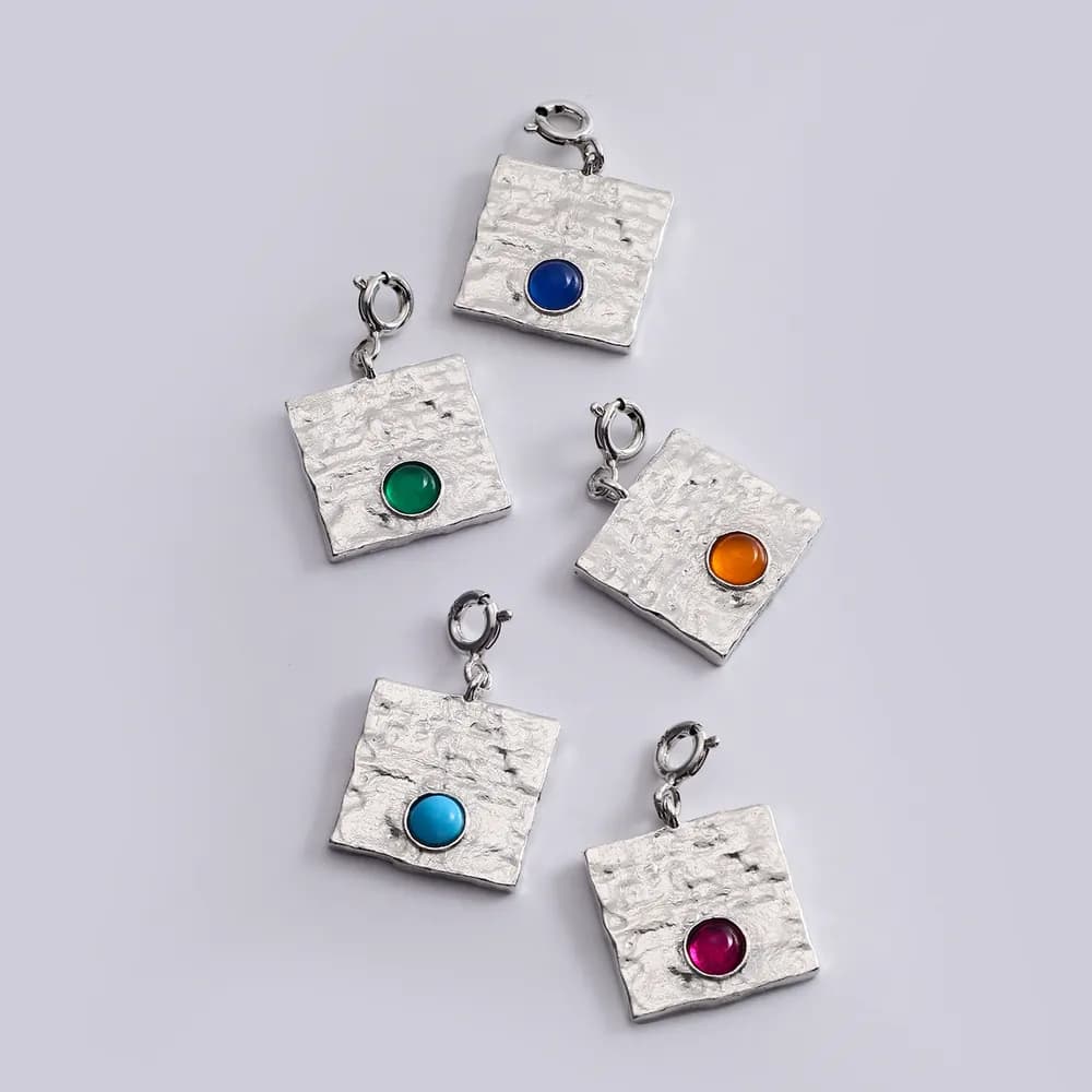 Textured square silver pendants each with a colored enamel dot in the center, arranged on a light background.