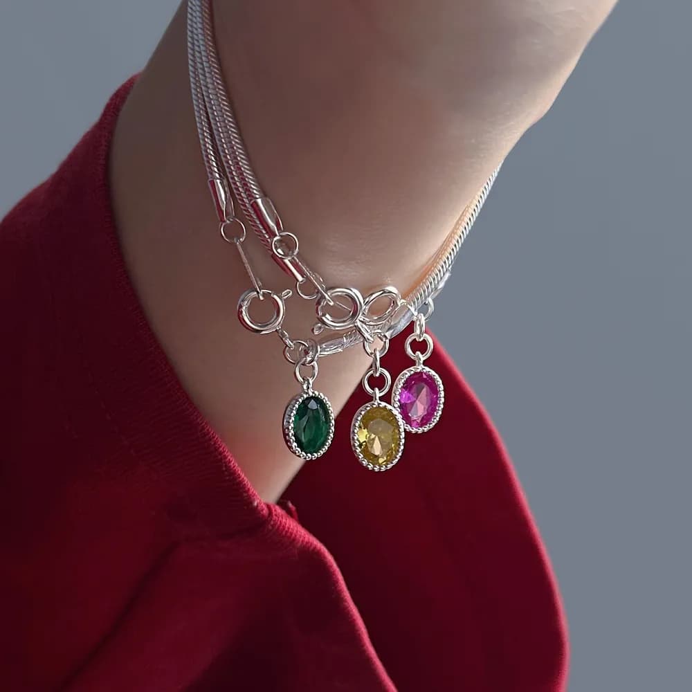 Close-up of a layered sterling silver bracelet with three oval gemstone charms in green, yellow, and pink against a red garment.