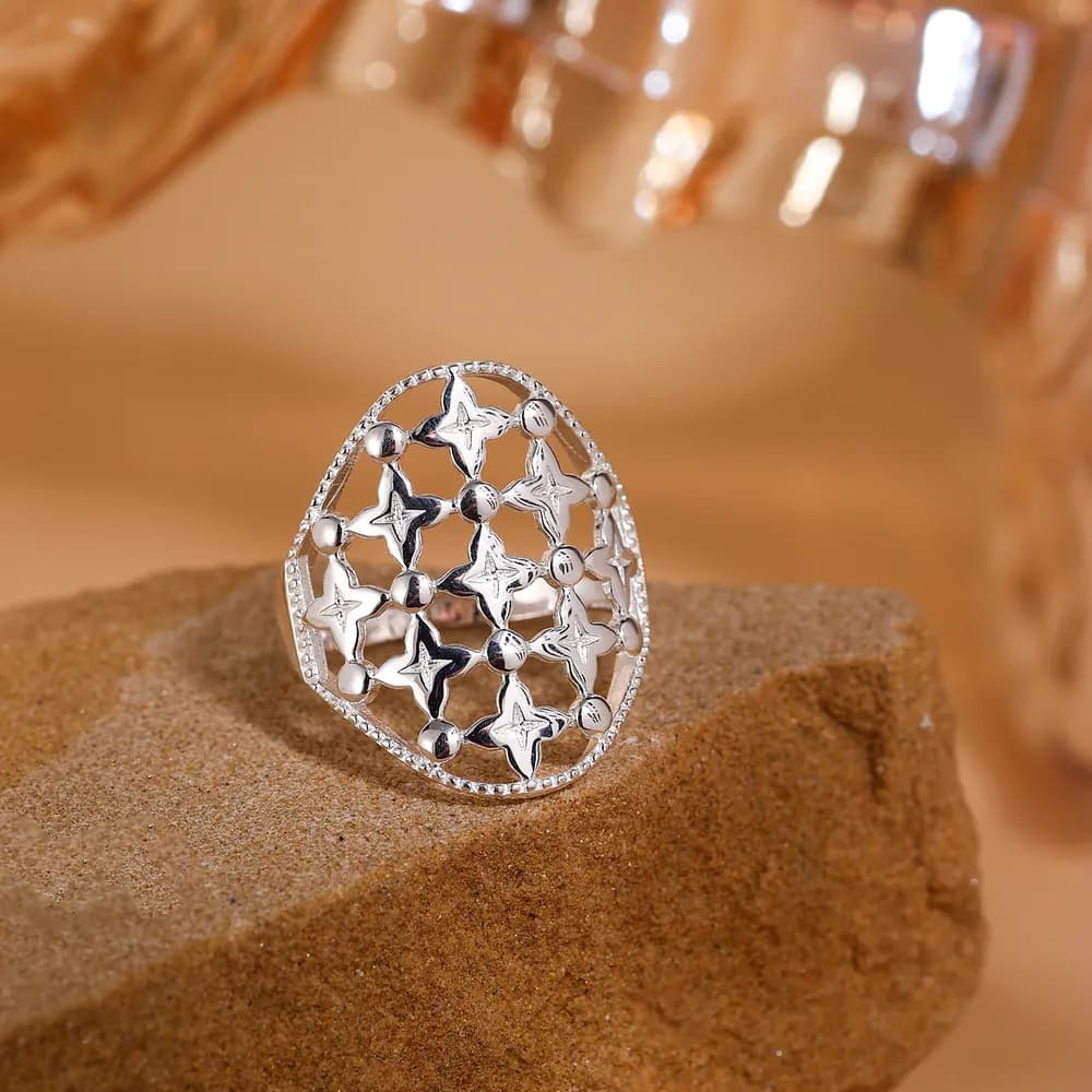A polished sterling silver ring with a circular star-lattice openwork design rests on sand.