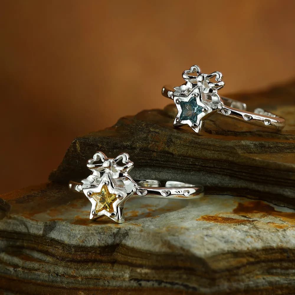 Two sterling silver rings with star-shaped settings and colored stones sit on layered stone slabs.