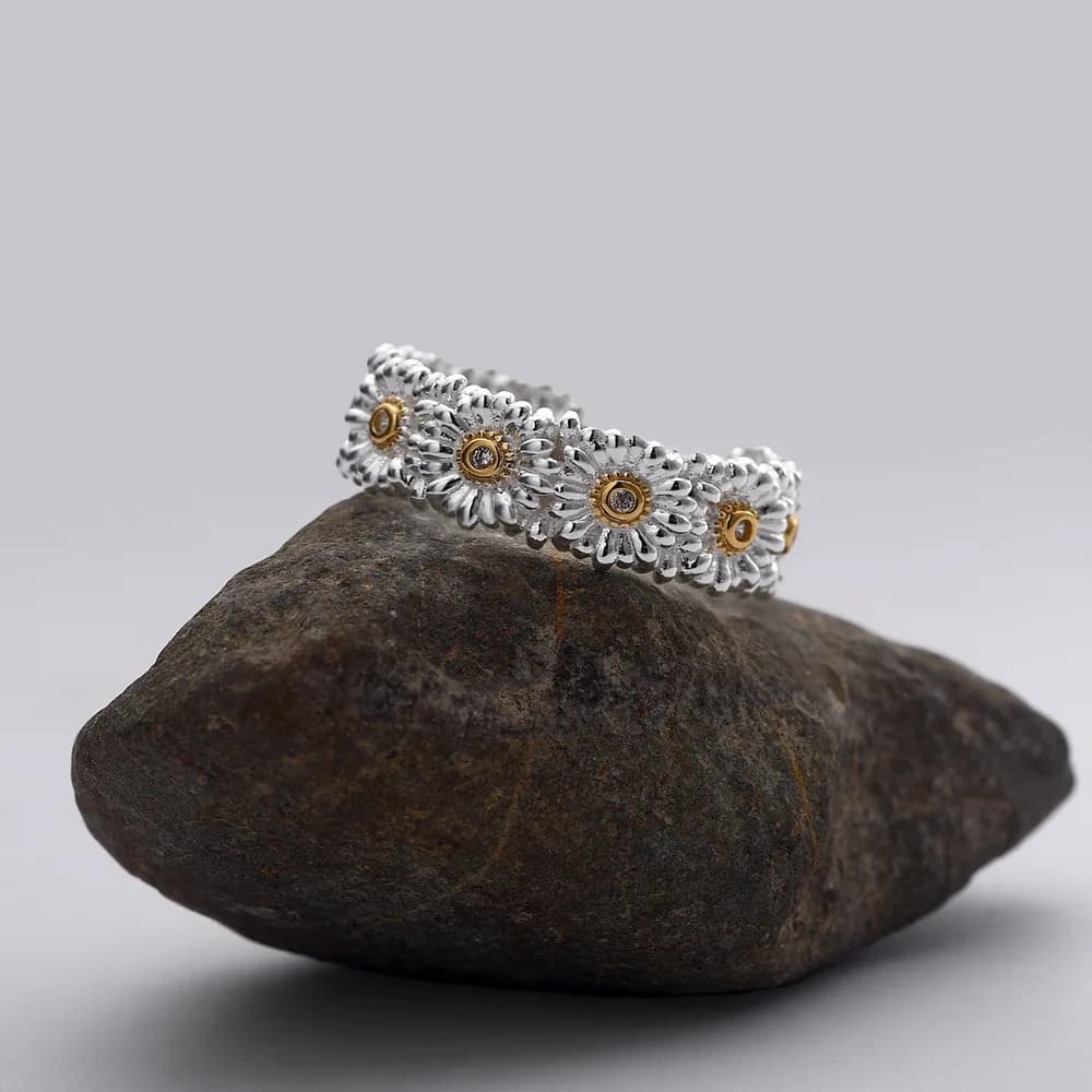Polished silver ring with a row of daisies and gold centers displayed on a dark rock