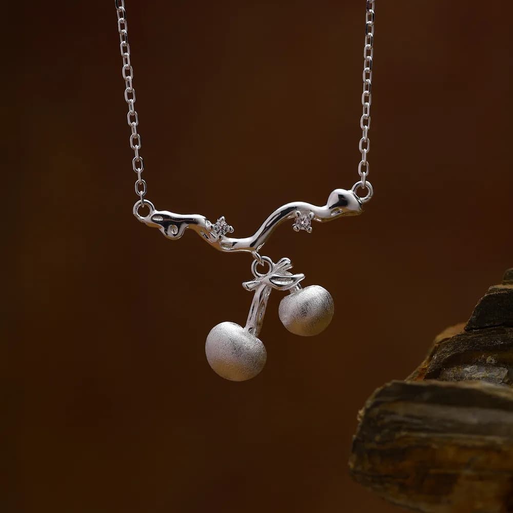 Polished sterling silver necklace with a curved branch pendant bearing two cherry-like beads and a small leaf detail.