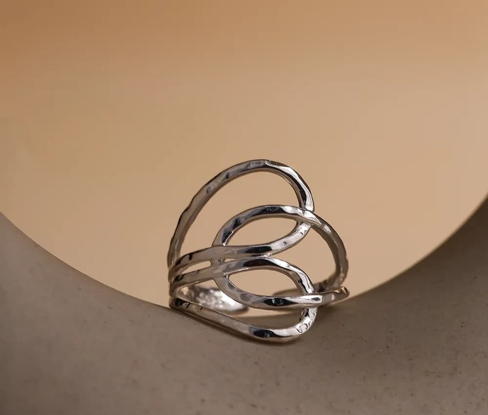 Polished sterling silver layered wave ring with interwoven bands resting on a curved beige surface.