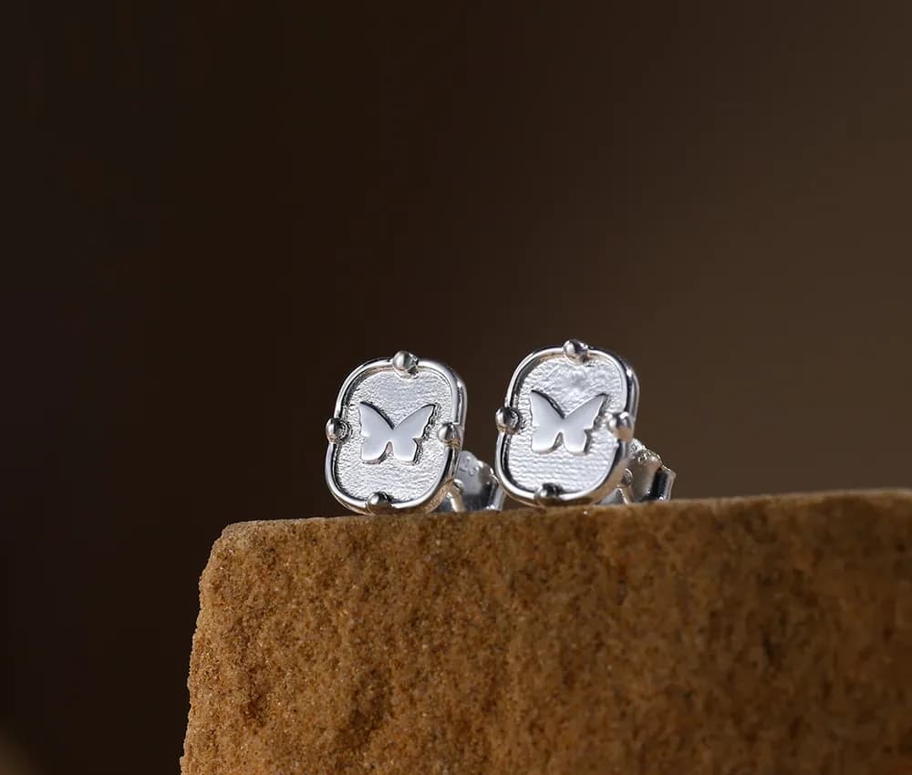 Close-up of silver square stud earrings with a butterfly cutout and dotted corner accents on a textured surface.