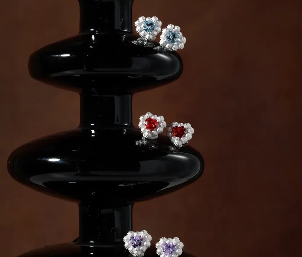 Three pairs of sterling silver stud earrings with heart-shaped colored stones surrounded by petal-like florets on a dark display.