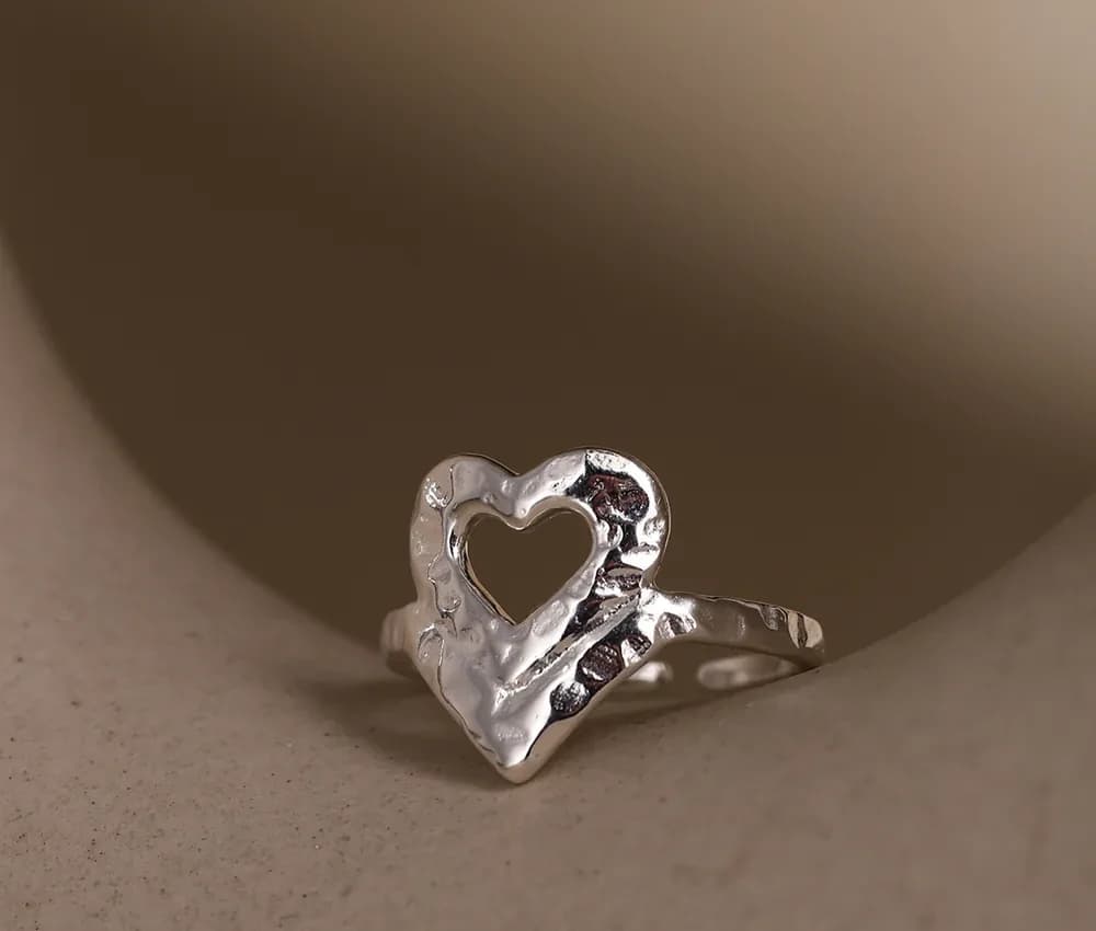 Polished sterling silver open-heart ring with a hammered texture on a soft, warm-toned surface.