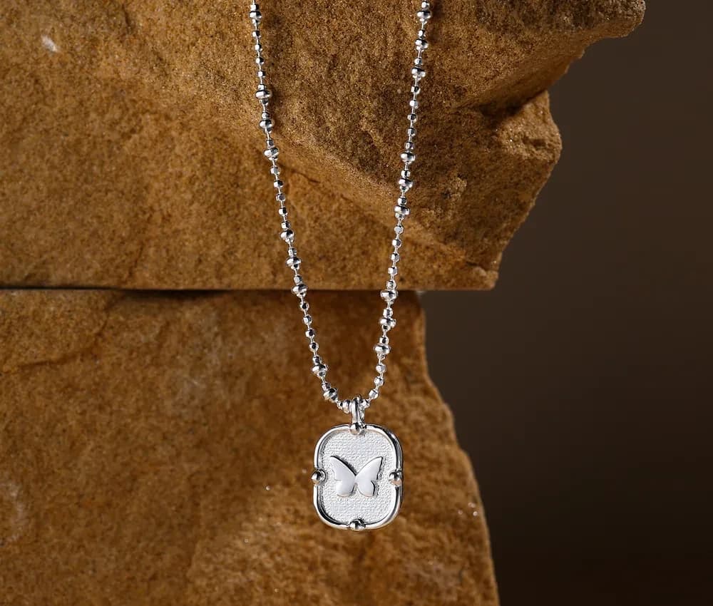 Polished silver pendant with a butterfly cut-out on a beaded chain necklace against a rocky backdrop.