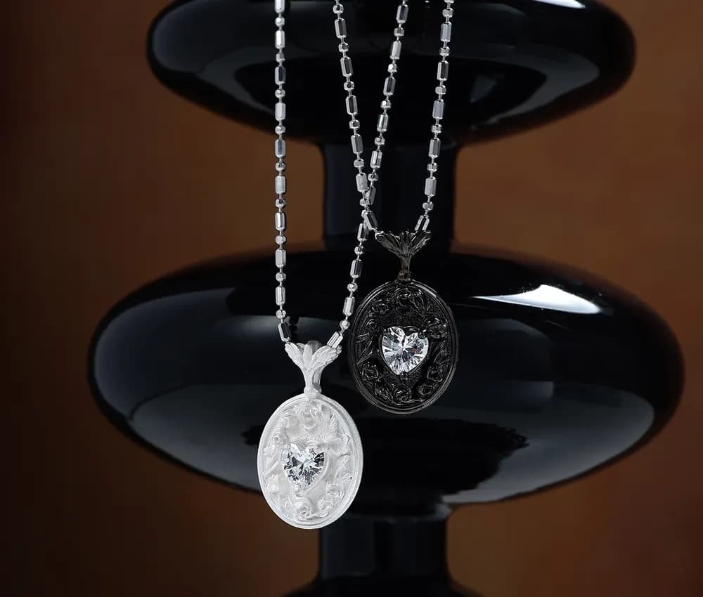 Two oval pendants with heart crystals on beaded chains, one polished silver and one oxidized with intricate filigree.