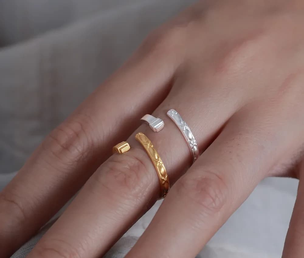 A hand wearing three polished rings: a gold faceted band, a silver faceted band, and a slim plain silver ring on the same finger.