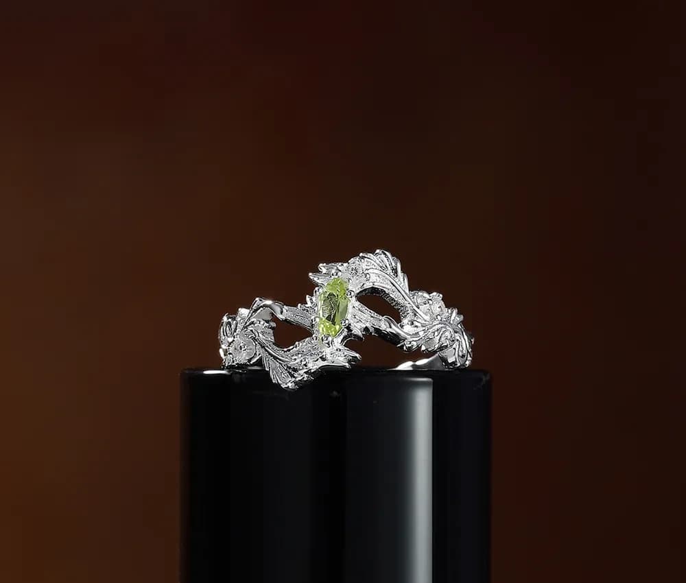Polished sterling silver ring with a leafy vine design and a central green gemstone displayed on a glossy black pedestal.