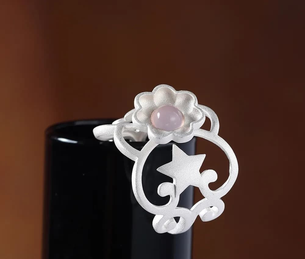 Sterling silver flower ring with a pink center sits on a dark pedestal.
