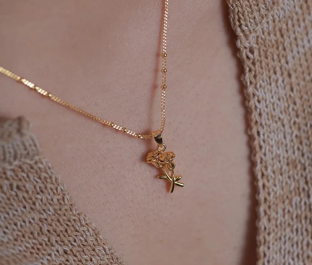Close-up of a delicate rose pendant necklace in polished sterling silver resting on skin and a knit garment.