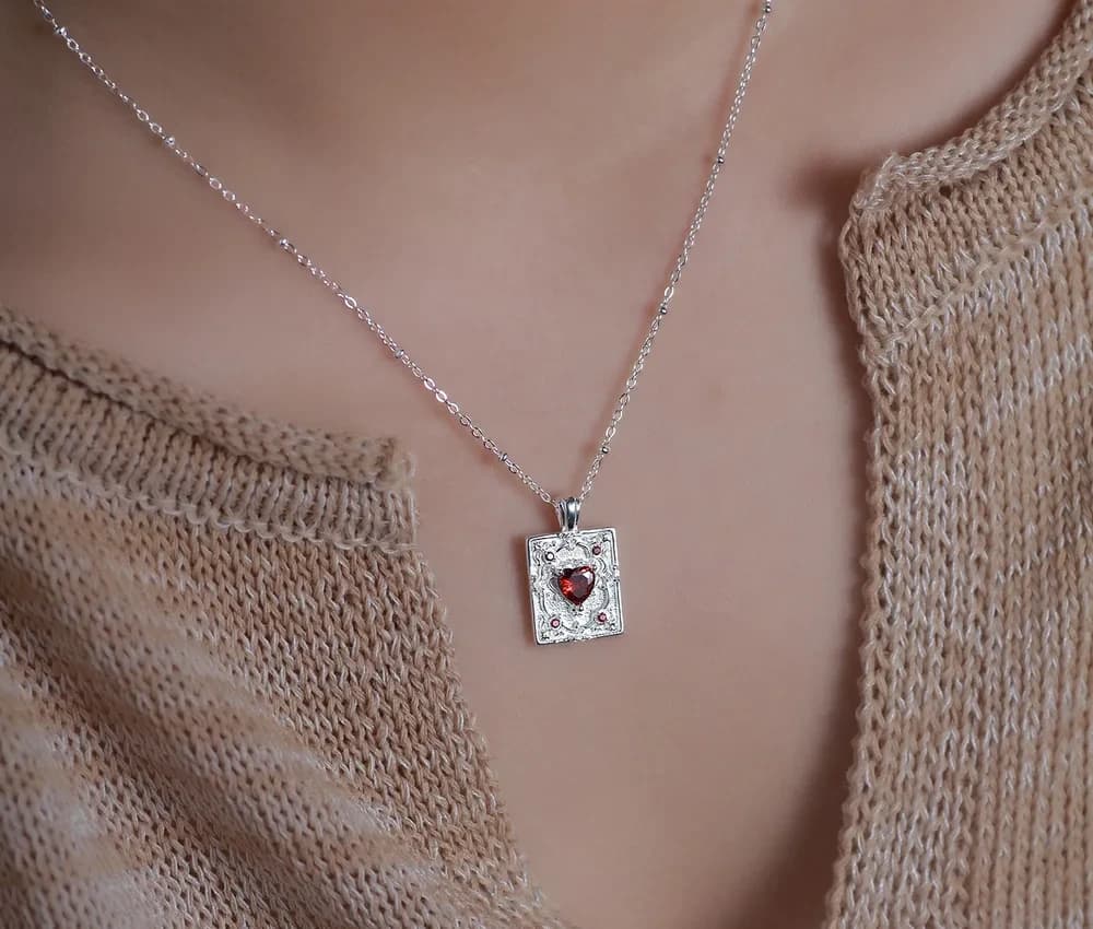 Delicate silver chain with a rectangular pendant featuring a heart-shaped red gem on the collarbone.