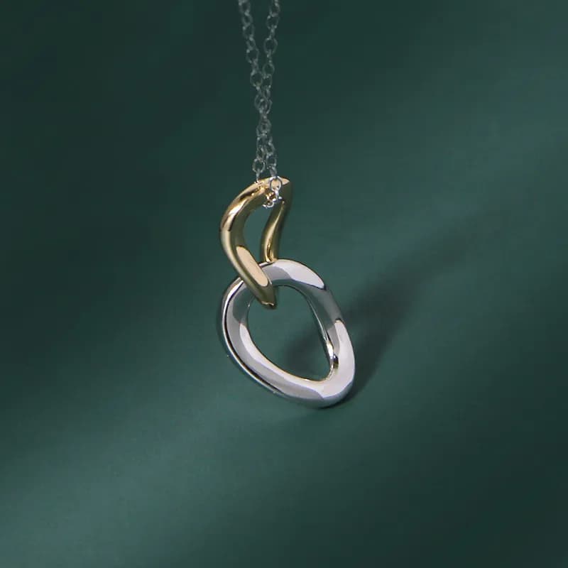 Two-tone interlocking rings pendant on a delicate chain against a dark green background.