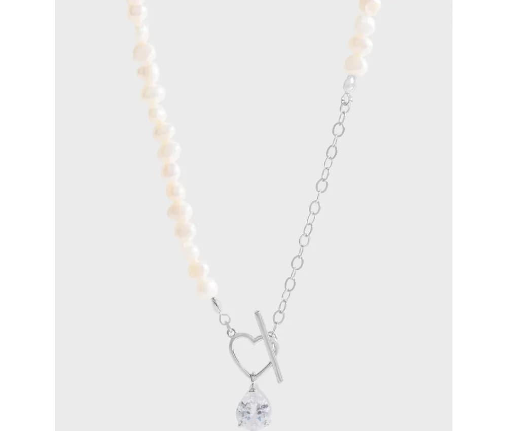 Silvery necklace with a heart toggle pendant and crystal drop, flanked by pearl and chain strands on a pale background.