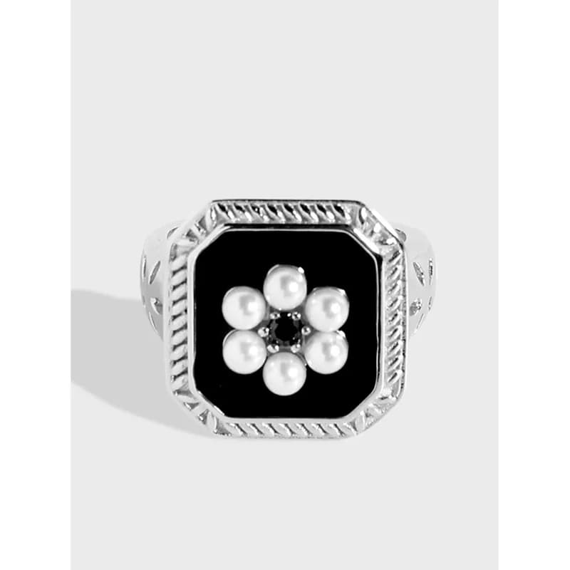Square silver ring with black enamel and a pearl flower centerpiece on a neutral background.