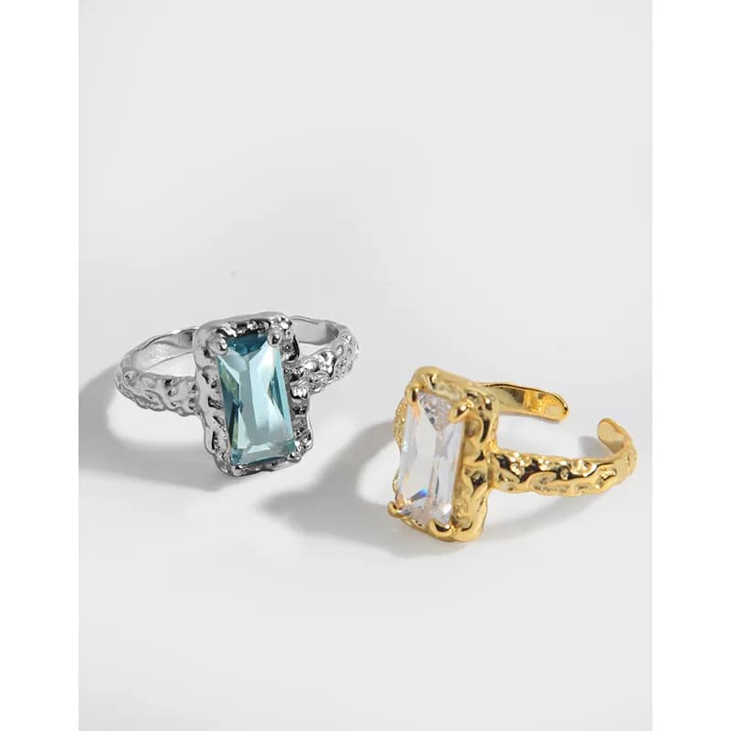 Two ornate rings with emerald-cut stones, one silver and one gold, shown side by side on a white background.