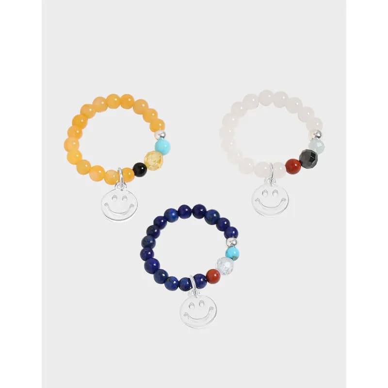 Three polished beaded rings with smile charms in yellow, white, and blue on a light background.