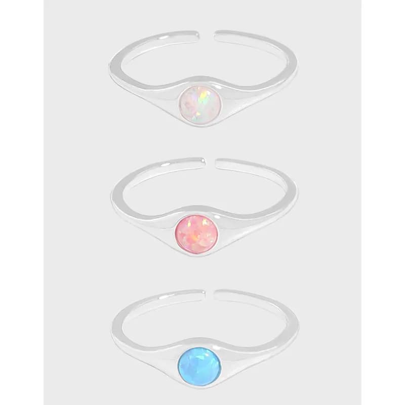 Three sterling silver rings with bezel-set round opal cabochons in pink, blue, and white on a light gray background.