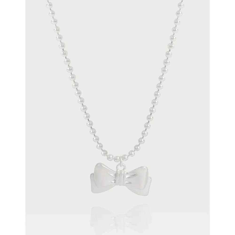 Polished silver bow pendant hanging from a beaded ball-chain necklace on a light backdrop.