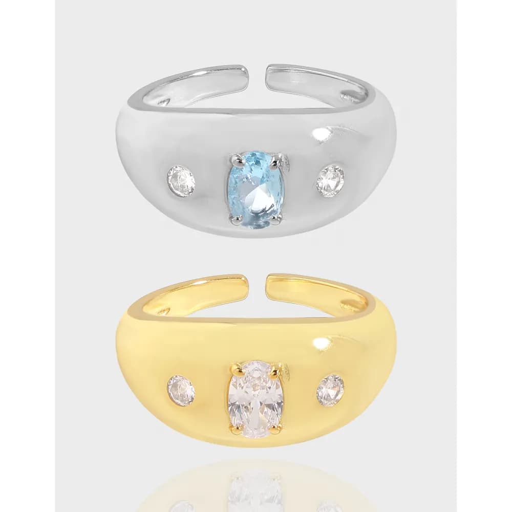 Two thick rings, one silver and one gold, each with an oval central gemstone flanked by two small stones on a smooth, rounded band.