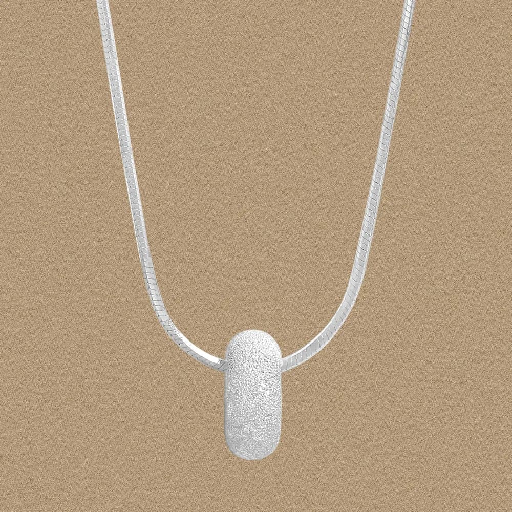 Close-up of a textured frosted cylinder pendant on a polished silver chain against a sandy background.