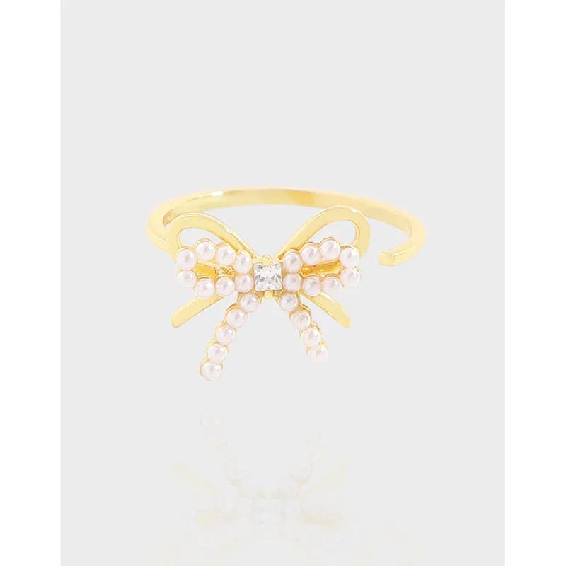 Gold-tone bow ring with pearl beads and a small central square gem on a slim polished band.