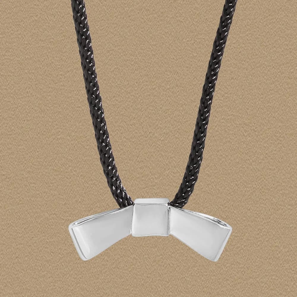 Polished sterling silver bow-tie pendant on a black braided necklace against a sandy background.