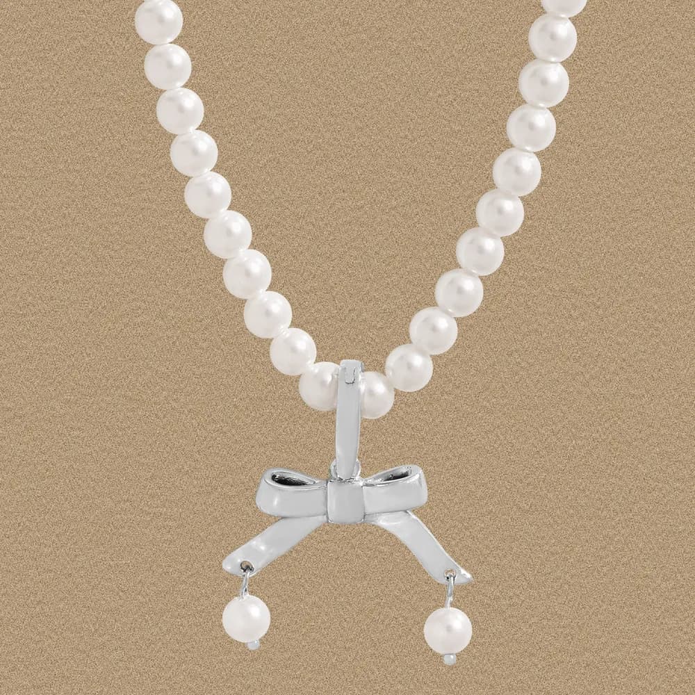 Pearl bead necklace with a silver bow pendant against a neutral beige background.