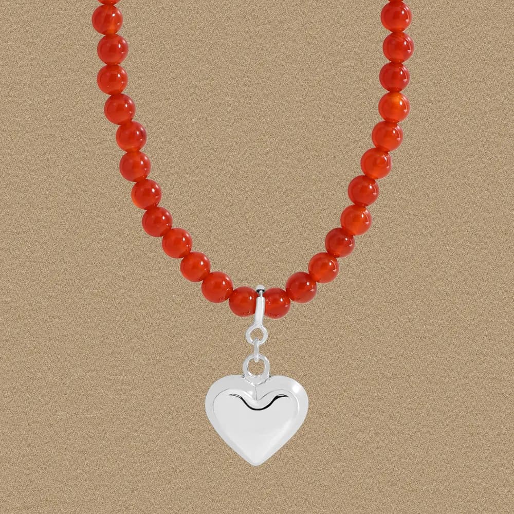 Close-up view of a red bead necklace with a silver heart pendant against a neutral backdrop.