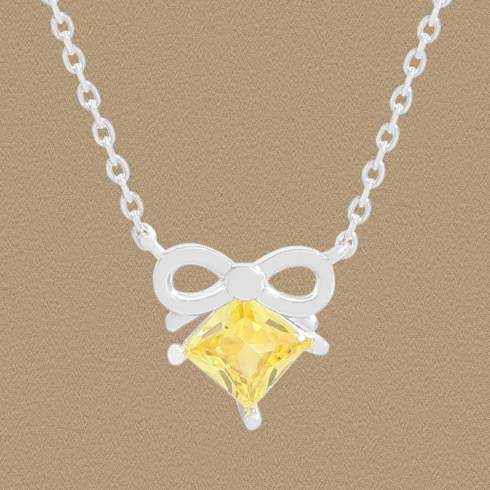 Silver bow pendant necklace with a yellow square-cut gemstone against a beige background.