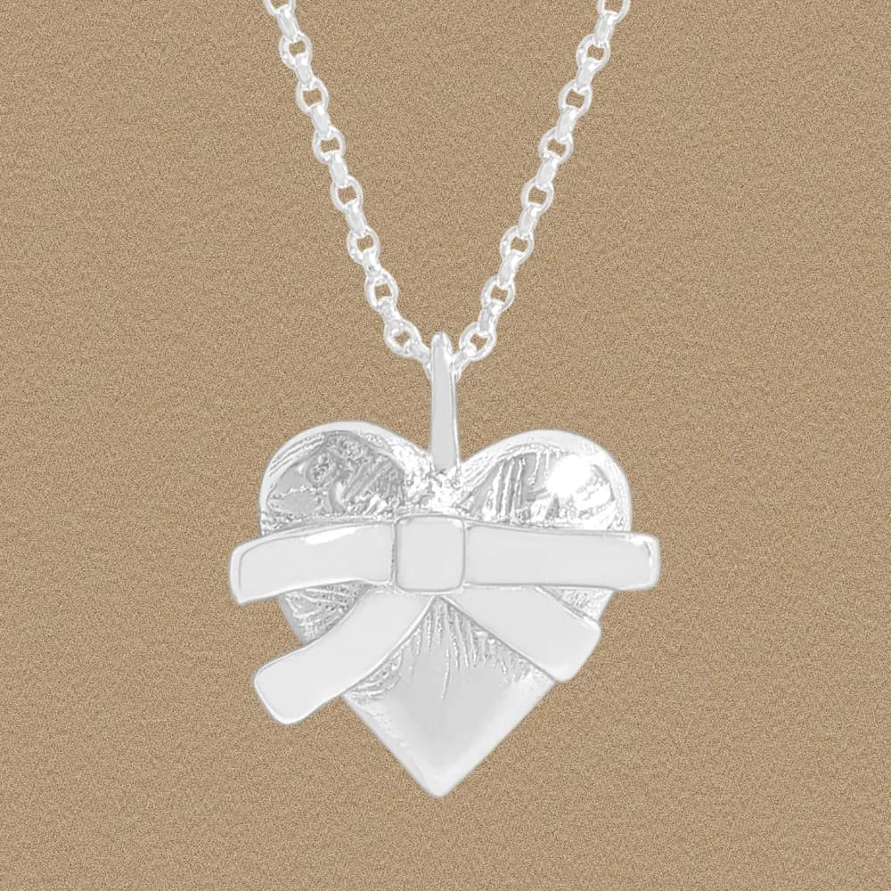 Polished sterling silver heart pendant with a bow on a chain against a warm beige background.