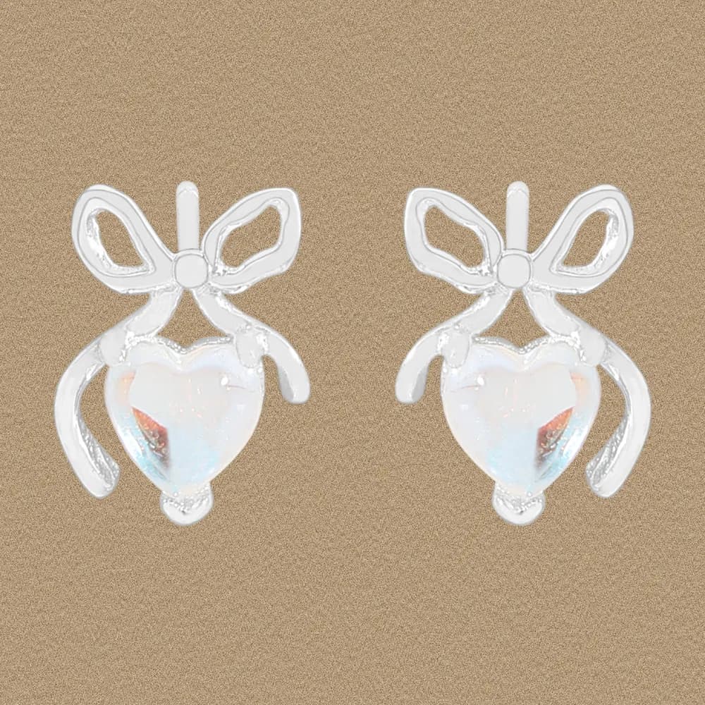 Polished sterling silver bow earrings with iridescent heart drops on a tan background.