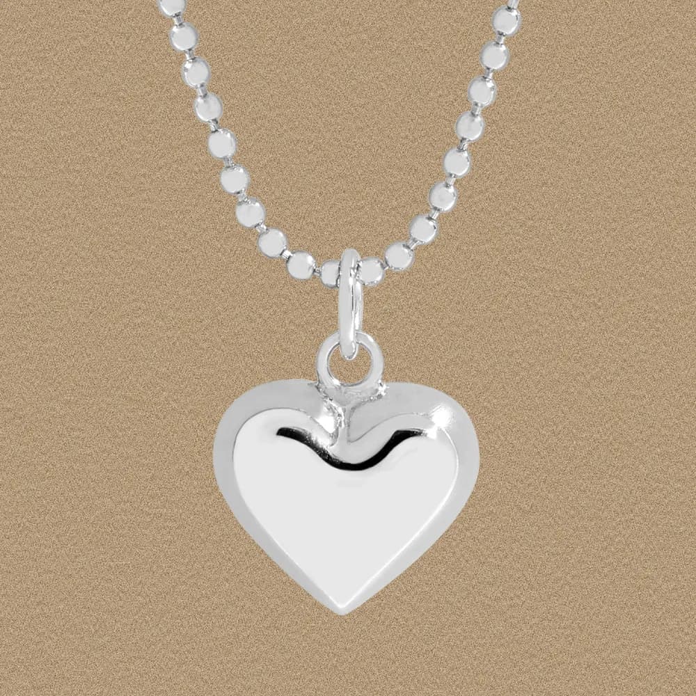Polished sterling silver heart pendant on a beaded chain necklace photographed against a beige backdrop.