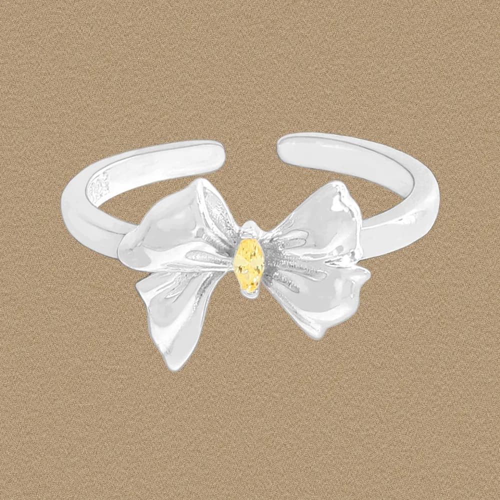 Polished sterling silver bow ring with a tiny yellow gemstone at the center on a beige background.