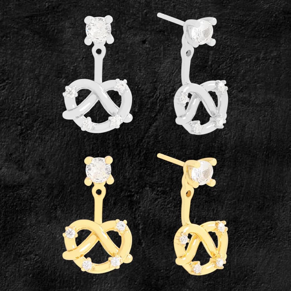 Polished infinity knot drop earrings in silver and gold-tone finishes with clear stones on a dark background.