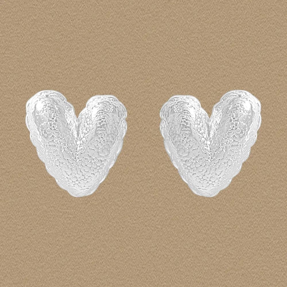 Pair of textured heart-shaped sterling silver stud earrings on a warm beige background.