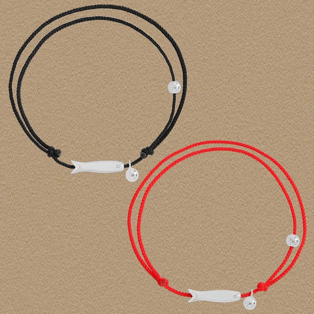 Two braided cord bracelets in black and red with a polished silver fish charm and a tiny disk charm on a sandy background.
