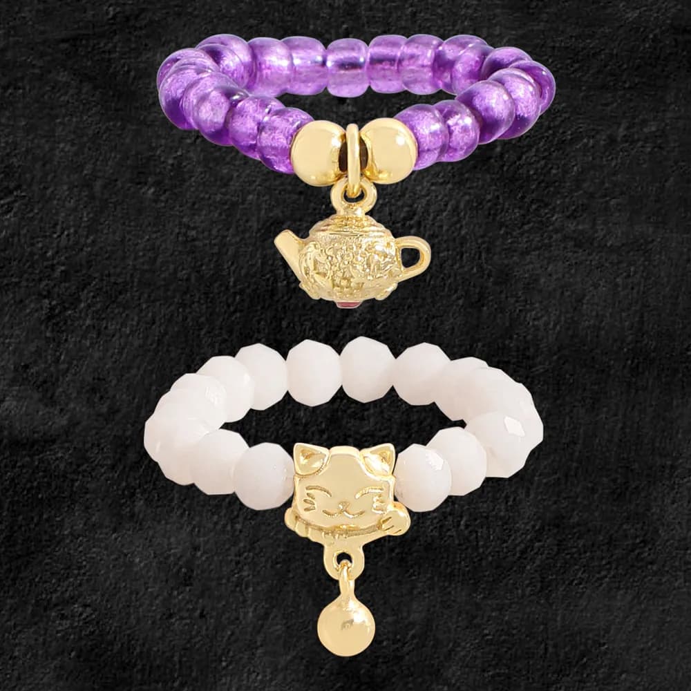 Two beaded stretch rings, one purple with a gold teapot charm, one white with a gold cat charm and dangling ball.