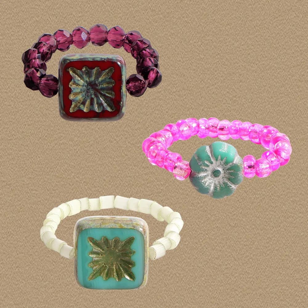 Three colorful square-bezel rings with sunburst center on a sandy background.