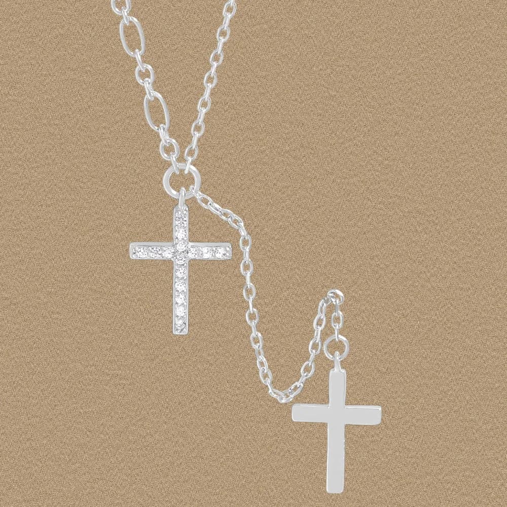 Two intertwined sterling silver chains with a pavé cross pendant and a plain cross pendant on a tan backdrop.