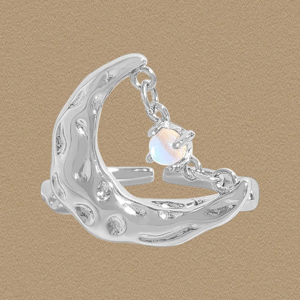 Polished silver crescent ring with a dangling opal charm on a tan background.