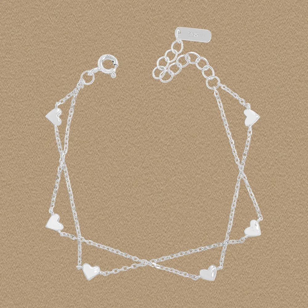 A polished sterling silver bracelet with two interwoven chains and small heart charms laid flat on a tan surface.