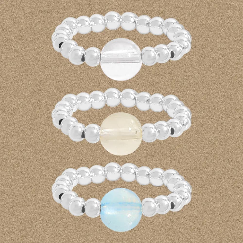 Three sterling silver beaded stretch rings with a large central bead in white, peach, and blue tones on a sand background.